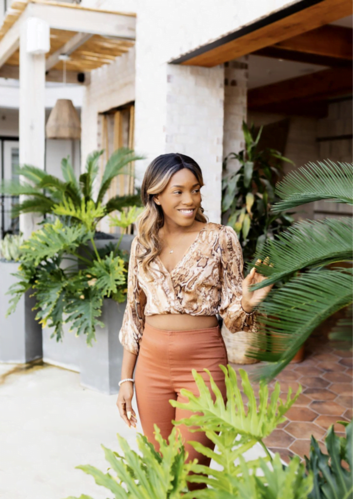 Destiny, a stunning black woman, walks out of a white stone entryway. She smiles as she touches a fern, looking over her left shoulder. She is wearing a brown-snake print cropped blouse and burnt orange pants.